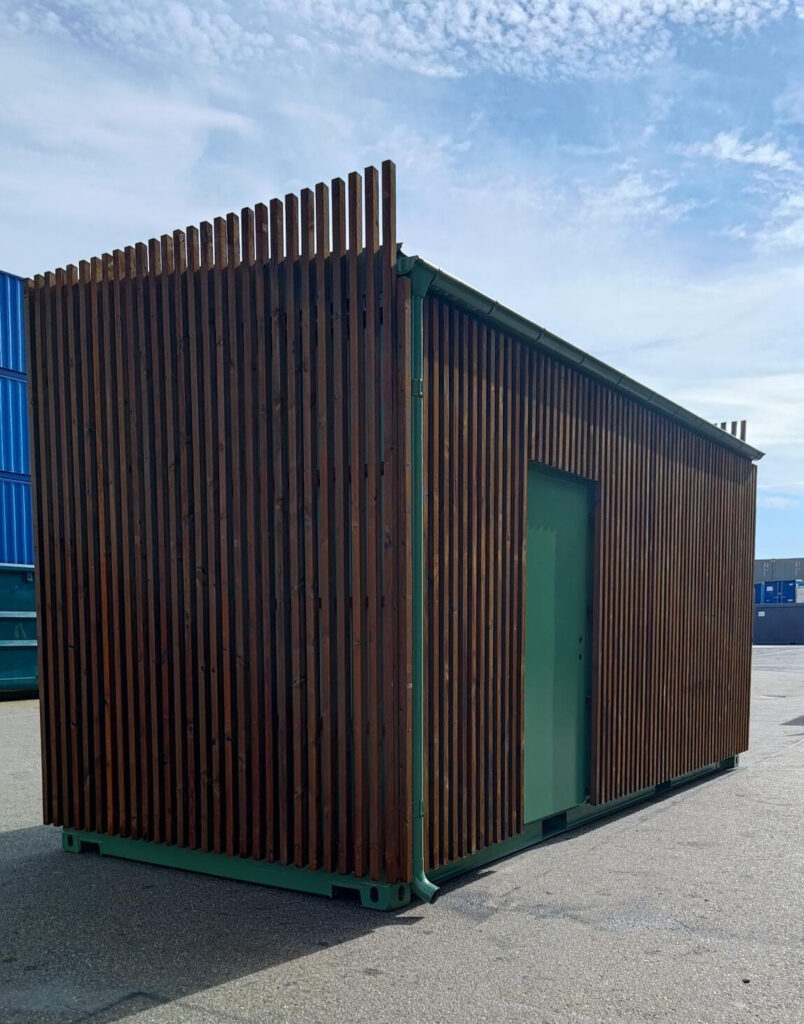 Mobil fjärrvärmestation byggd av container, klädd med vertikala träpaneler - vy snett framifrån.