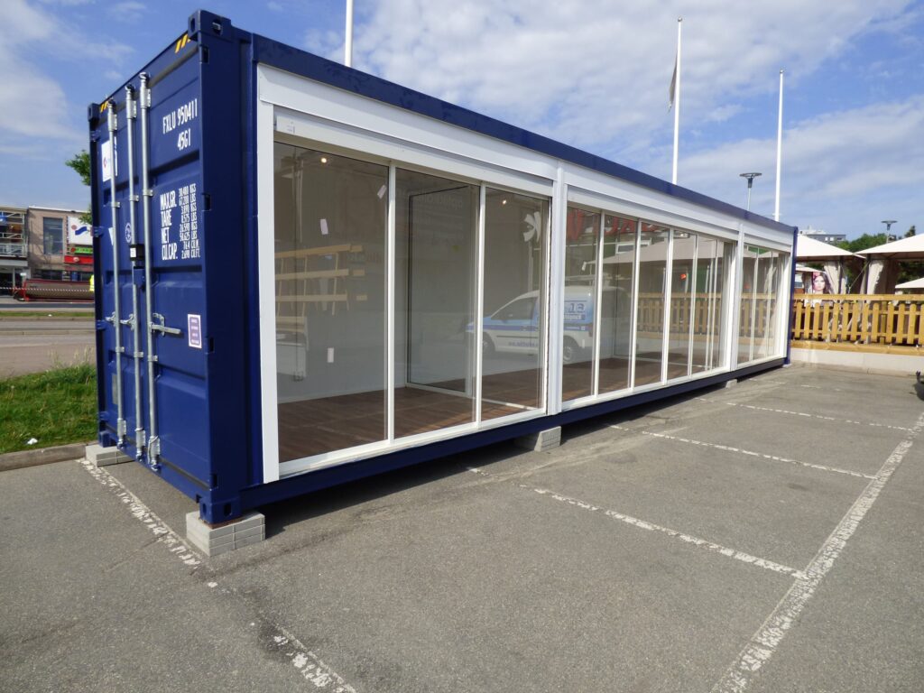Blå butikscontainer med glasfasad och skjutdörrar på parkeringsplats, byggd av LCS
