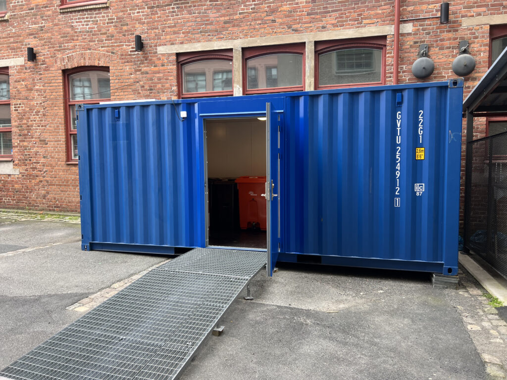 Frontvy på miljörumscontainer med öppen gångdörr och ramp för tillgänglighet, byggd av LCS