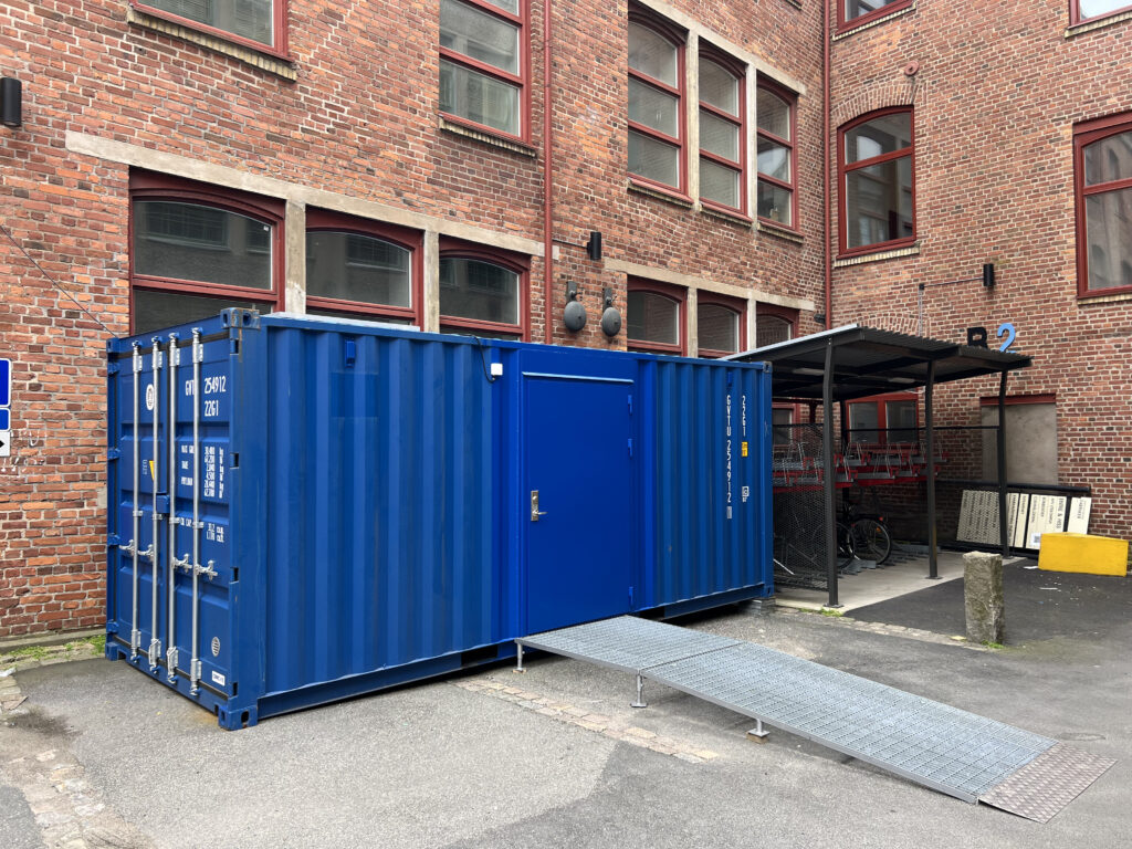 Sidovy på miljörumscontainer med gångdörr och ramp i urban miljö, byggd av LCS