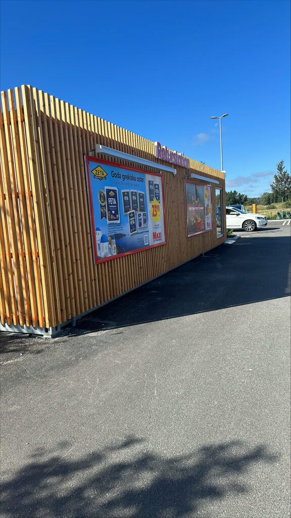 Långsida på pantstationscontainer med träfasad och reklamskyltar, byggd av LCS för matbutik